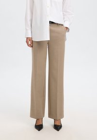 Beežid laiad püksid, mis on valmistatud siledast kangast, omades kahte külgtaskut, voldijoont ja kohandatud sobivust vöökohtadel. Mustad kontsad.