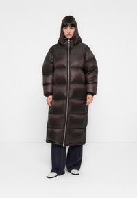 Vrouw in een lange, donkerbruin gewatteerde jas met capuchon, zwarte wijde broek en witte schoenen, staande tegen een effen witte achtergrond.