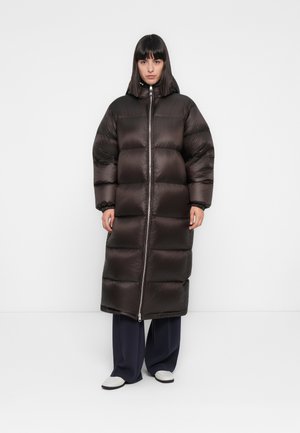 Vrouw in een lange, donkerbruin gewatteerde jas met capuchon, zwarte wijde broek en witte schoenen, staande tegen een effen witte achtergrond.