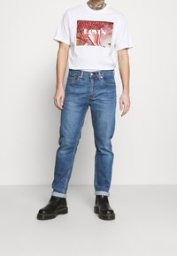 Vit t-shirt med en färgglad Levi's grafik, ihop med blå denimjeans och svarta ankelskor. Avslappnad stil, ledig passform.