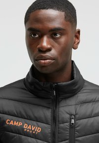 Chaqueta acolchada negra con cuello alto, con textura acolchada y un bolsillo con cremallera en ángulo. Logo "CAMP DAVID SPORT" en naranja en el pecho.