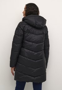 Manteau matelassé noir avec capuche, présentant un motif en chevrons, un design matelassé et une texture lisse. Comprend un détail de logo sur la manche.
