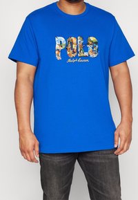 Blå bomullströja med en färgglad "Polo Ralph Lauren" grafisk design på framsidan, med korta ärmar och rundad halsringning.