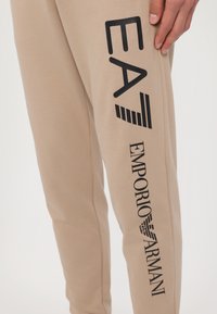 Beige joggebukser laget av bomullsblanding, med trykte svarte logoer "EA7" og "Emporio Armani" på siden. Glatt tekstur, tapered design.