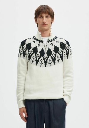 Un homme porte un pull crème à demi-zip, orné d’un empiècement géométrique noir marqué aux épaules, avec un pantalon foncé, sur un fond blanc uni.