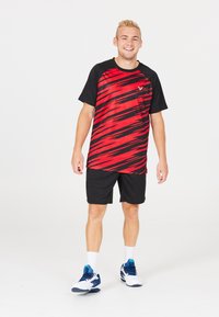 Rød og sort kortærmet sportstrøje med et stribet mønster, parret med sorte shorts og hvide sneakers med blå accenter.