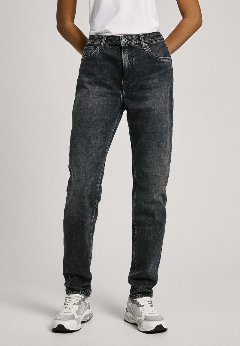 Vue de face d'une personne portant un jean noir délavé taille haute, des baskets blanches et un haut blanc sans manches, les mains dans le dos.