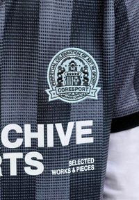 Schwarz-grau kariertes Stoff mit einem weißen, gestickten Emblem mit der Aufschrift "Perpetual Celebration of Art in Sport" sowie gedrucktem Text "CHIVE" und "Selected Works & Pieces".