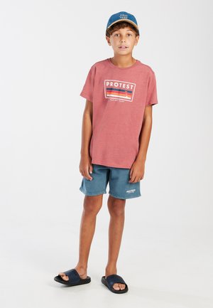 Rode katoenen T-shirt met "PROTEST" logo, blauwe shorts en zwarte slippers. Jongeman staat tegen een effen witte achtergrond.