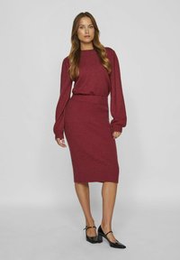 Set din tricot burgund cu două piese, format dintr-o bluză cu mâneci lungi și guler rotund și o fustă tip creion cu dungi. Asortat cu pantofi cu toc pătrat negri.