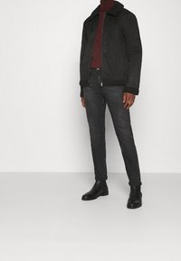 Schwarze Bomberjacke aus Wildleder mit einem Lammfellkragen, kombiniert mit dunklen Jeans und schwarzen Stiefeletten. Schlichtes, zeitgemäßes Design.