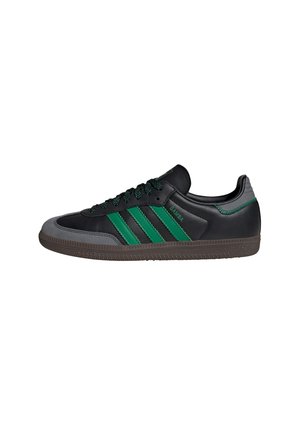 Black leather sneaker with green accents, featuring three stripes, textured gray toe cap, and black laces. Rubber sole with traction pattern.