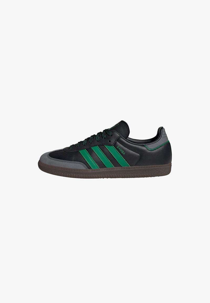 Black leather sneaker with green accents, featuring three stripes, textured gray toe cap, and black laces. Rubber sole with traction pattern.