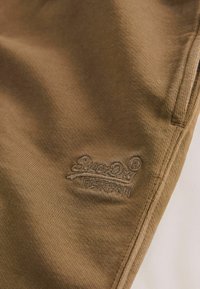 Gros plan sur un tissu marron avec un logo SuperDry cousu près de la couture d’une poche latérale.