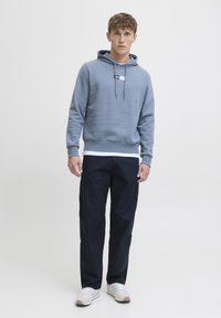 Uomo che indossa una felpa blu con cappuccio, una camicia bianca sotto, pantaloni scuri e sneakers bianche, in piedi davanti a uno sfondo chiaro e semplice.