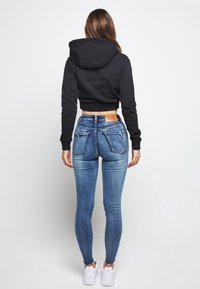 Mujer de espaldas con un hoodie negro corto, jeans ajustados azules desgastados y zapatillas blancas frente a un fondo blanco liso.