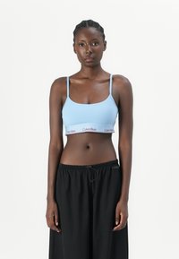 Lichtblauwe bralette met dunne bandjes en elastische band met daarop het rode 'Calvin Klein' logo; gecombineerd met losse zwarte broek. Zachte textuur.