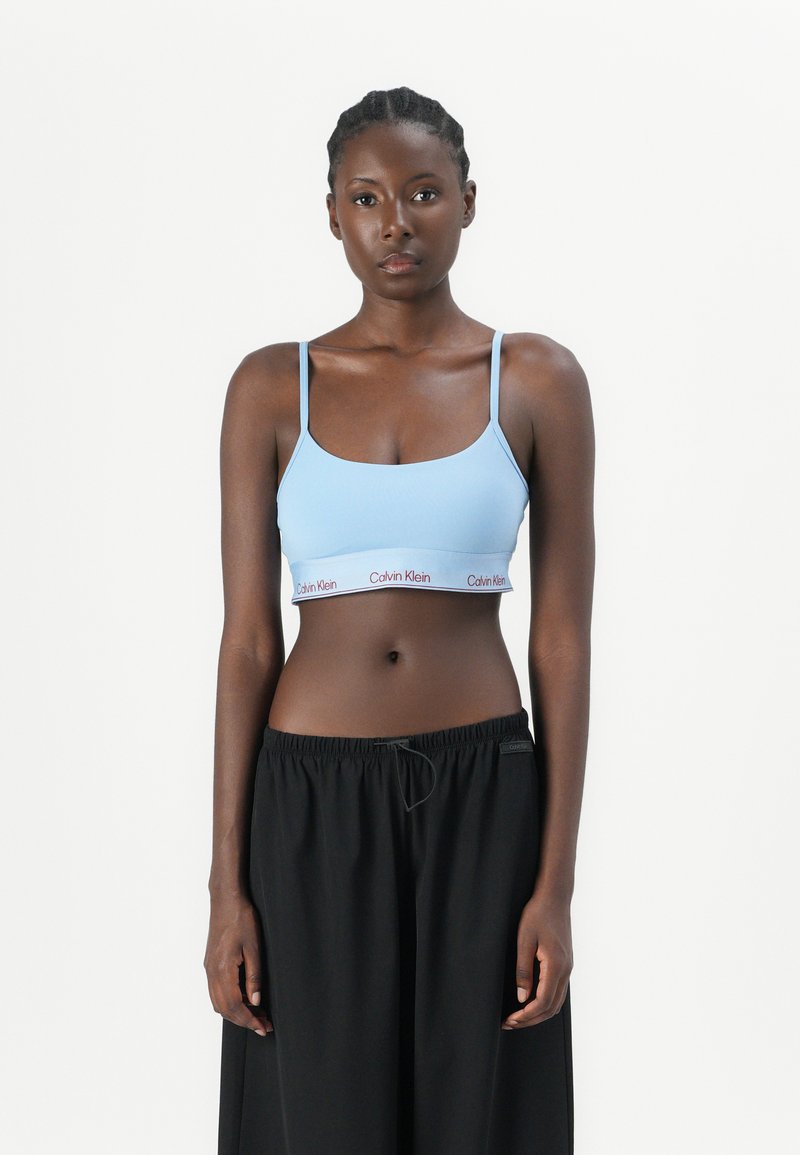 Lichtblauwe bralette met dunne bandjes en elastische band met daarop het rode 'Calvin Klein' logo; gecombineerd met losse zwarte broek. Zachte textuur.