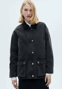 Black denim jacket with a collar, front snap closures, and two side pockets. Features a relaxed fit and visible stitching details.
