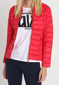 Červená vyp padded bunda s předním zipem, bočními kapsami, těsným střihem a hladkým, lesklým povrchem. Nositá přes bílou košili.