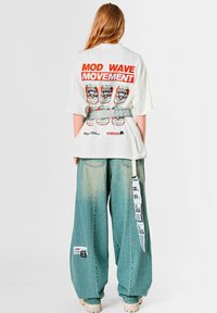 Camiseta blanca de gran tamaño con texto gráfico rojo y diseños de latas coloridas en la parte trasera; combinada con pantalones vaqueros de mezclilla de gran tamaño en azul claro con parches.