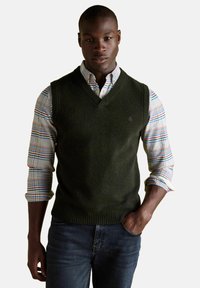 Černoch v tmavozelenej pletenej veste cez viacfarebnú kockovanú košeľu s vyhrnutými rukávmi a tmavými džínsami, s jednou rukou v vrecku.