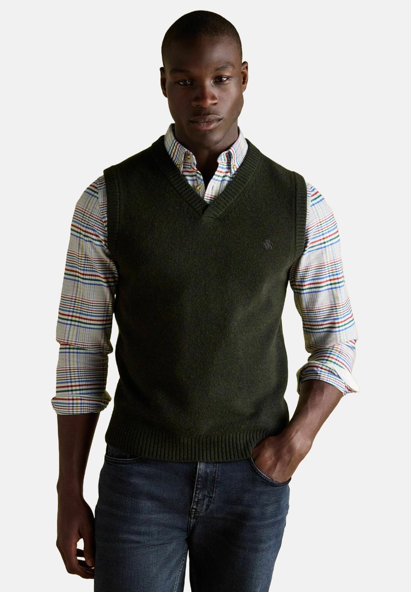 Černoch v tmavozelenej pletenej veste cez viacfarebnú kockovanú košeľu s vyhrnutými rukávmi a tmavými džínsami, s jednou rukou v vrecku.