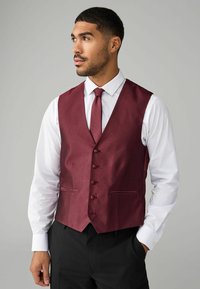 Burgundský vest s textúrovaným vzorom, šesť gombíkov a dve predné vrecká, nosený na bielom tričku a doplnený zodpovedajúcou kravatou.