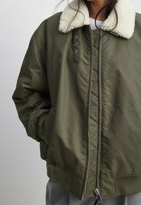 Chaqueta bomber verde oliva con cuello de piel sintética crema, cierre de cremallera y bolsillos laterales. Presenta detalles acolchados y bajo acanalado.