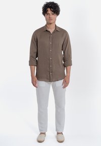 Camicia button-up in lino marrone con maniche arrotolate, abbinata a pantaloni beige chiaro e espadrillas neutre. Design semplice e pulito.
