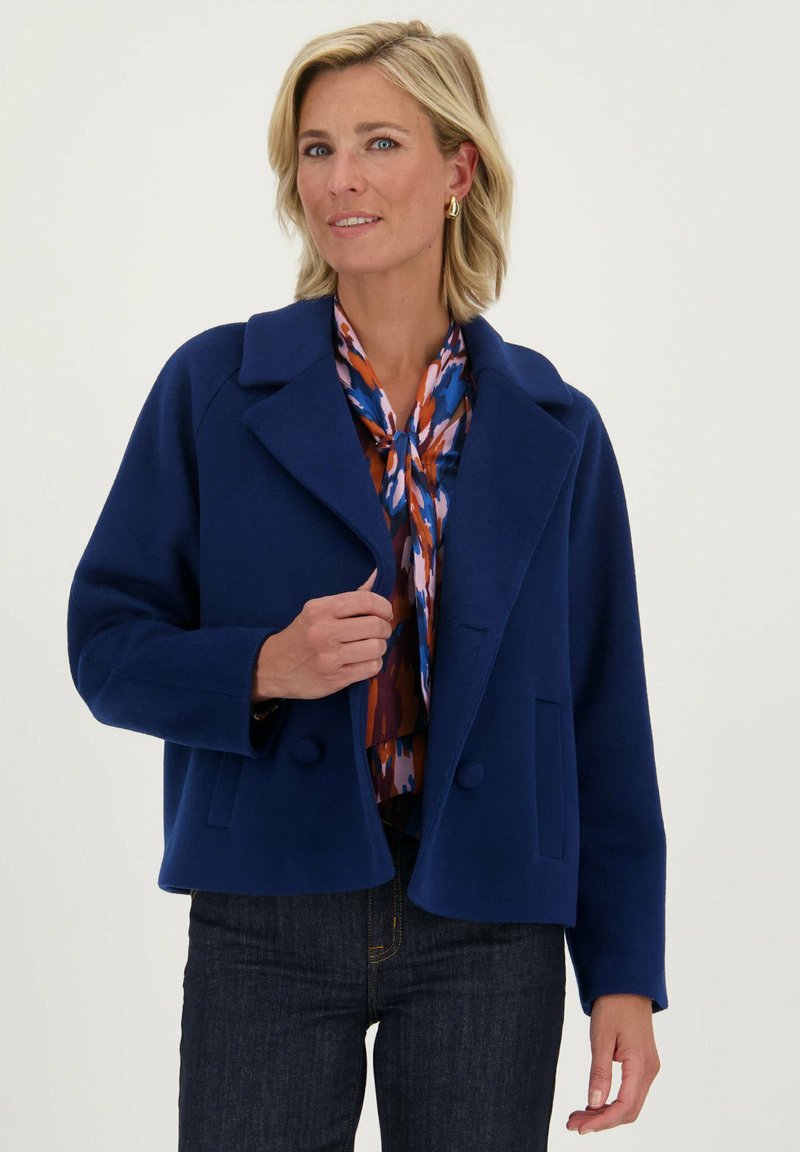Marineblauwe cropped jas van wol, met een grote revers, dubbele knoopsluiting en voorzakken. Draagt over een gebloemde blouse.