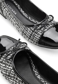 Ballerine in bianco e nero con tomaia in tweed tessurizzato, dettagli in vernice lucida e un fiocco decorativo sulla punta.