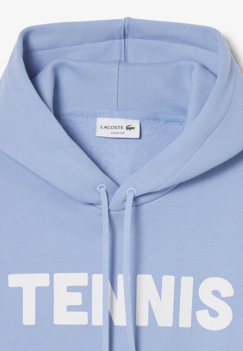 Felpa con cappuccio azzurro chiaro con scritta bianca "TENNIS" sul davanti, coulisse al collo e un'etichetta Lacoste all'interno del colletto con la dicitura "Loose Fit".