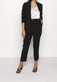 Blazer noir à manches longues porté sur un caraco en satin blanc, accompagné d'un pantalon noir ajusté et de sandales à talons noires à brides.