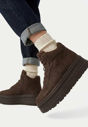 Brązowe sneakersy na platformie z wysoką cholewką i kwiatowym haftem, noszone z kremowymi prążkowanymi skarpetkami i podwiniętymi ciemnymi dżinsami na jasnym tle.