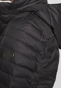 Giacca a vento nera con texture trapuntata, spalline ben pronunciate e un logo circolare sulla manica. Dotata di chiusura con zip.