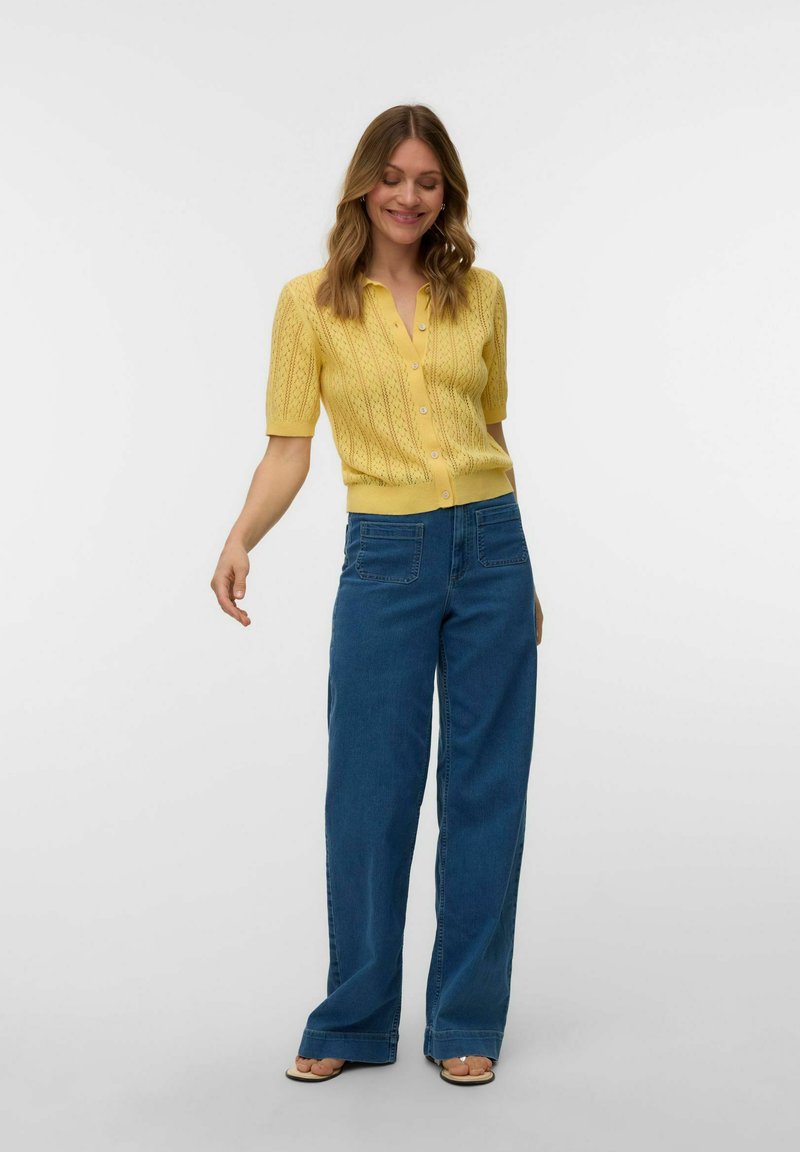 Femme portant un cardigan jaune à manches courtes boutonné et un jean bleu à jambes larges, debout avec un léger sourire et une posture détendue.