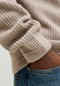 Beżowy sweter dzianinowy z prążkowanymi mankietami, lekko złożony. Zbliżenie ukazuje miękką fakturę i szczegóły dzianiny, zestawione z niebieskimi dżinsami.