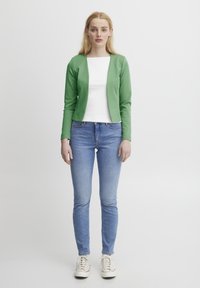 Zelený dlouhý svetr s rukávy, bílý přiléhavý top, světle modré skinny džíny a béžové tenisky. Jednoduchý design; žádné viditelné vzory nebo kovové prvky.