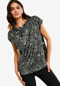 O femeie cu păr închis la culoare poartă un top negru cu mânecă scurtă acoperit cu paiete argintii strălucitoare și pantaloni negri, pozând pe un fundal deschis.