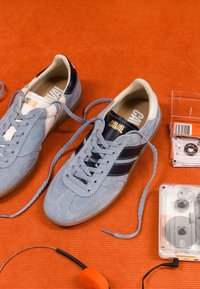 Paire de baskets en daim bleu clair avec des bandes noires et des lacets défaits, posée sur un tissu orange à côté de cassettes transparentes et d’un casque orange.