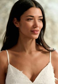 Top alb cu bretele subțiri, cu detalii texturate. Subiectul are păr lung și închis la culoare, iar fundalul este neutru. Tonalitatea pielii pare caldă.