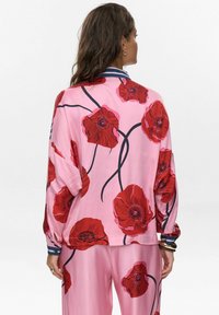 Blouse à manches longues en tissu rose doux, ornée d'un grand imprimé floral de coquelicots rouges, avec des accents de tige foncée et des détails de col rayé.