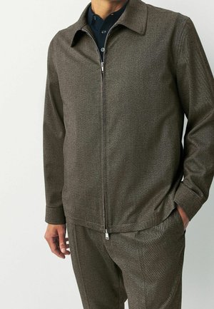 Homme portant une veste marron zippée avec un col par-dessus une chemise bleu foncé, une main dans la poche et l'autre détendue le long du corps.