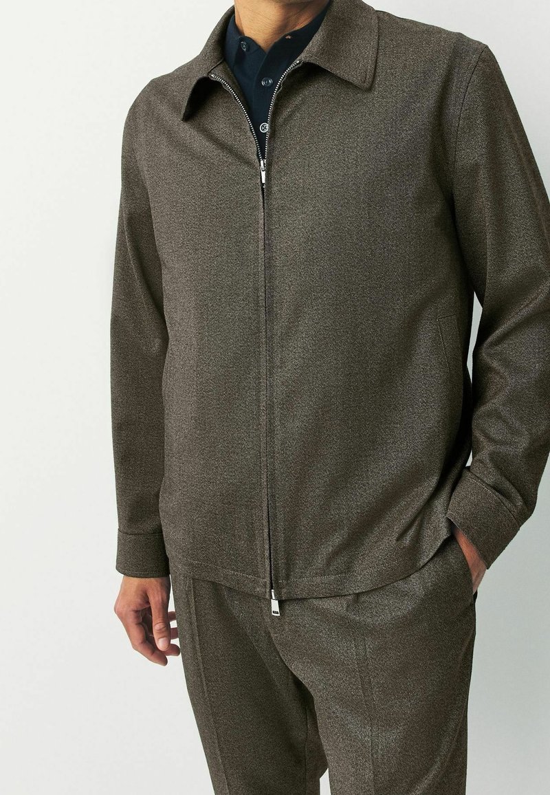 Homme portant une veste marron zippée avec un col par-dessus une chemise bleu foncé, une main dans la poche et l'autre détendue le long du corps.