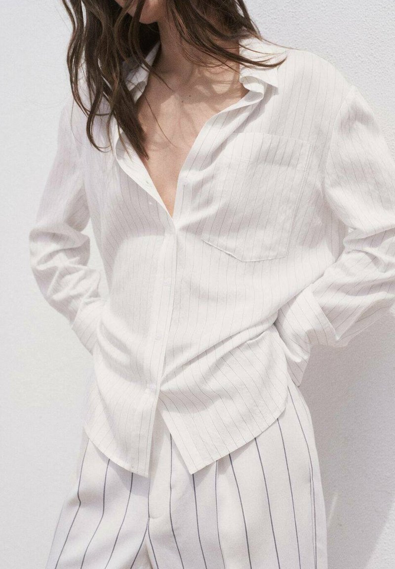 White, long-sleeve shirt with vertical pinstripes, a pocket, and a relaxed collar. Worn with matching striped trousers. Soft, lightweight fabric.