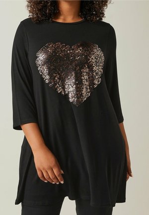 Femme portant une tunique noire à manches trois quarts avec un grand cœur pailleté sur la poitrine.