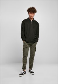 Fekete, hosszú ujjú póló fél cipzárral, olíva zöld kockás cargo nadrág és fekete edzőcipő fehér fűzőkkel. Lazán illeszkedő outfit.