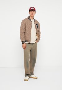 Brun rutete bomberjakke med marineblå kant og brodert "P"-logo. Kombinert med en lys beige t-skjorte, beige bukser og hvite sneakers.