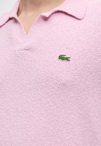 Κοντινό πλάνο σε ένα ροζ πικέ πόλο με μικρό πράσινο λογότυπο κροκόδειλου στην αριστερή πλευρά του στήθους.